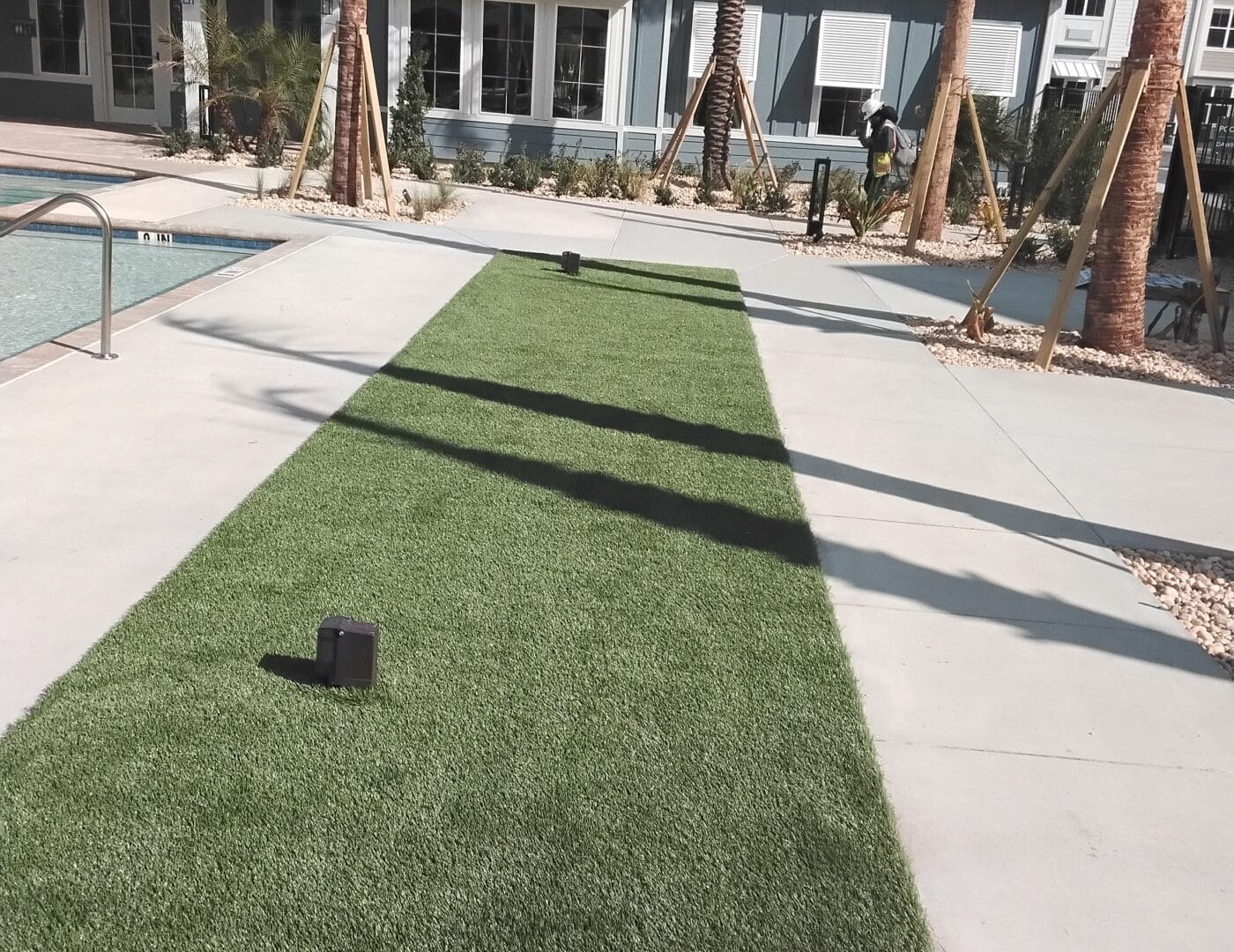 A long strip of artificial grass with small black lights on both sides lines a pool, bordered by palm trees and a modern building.