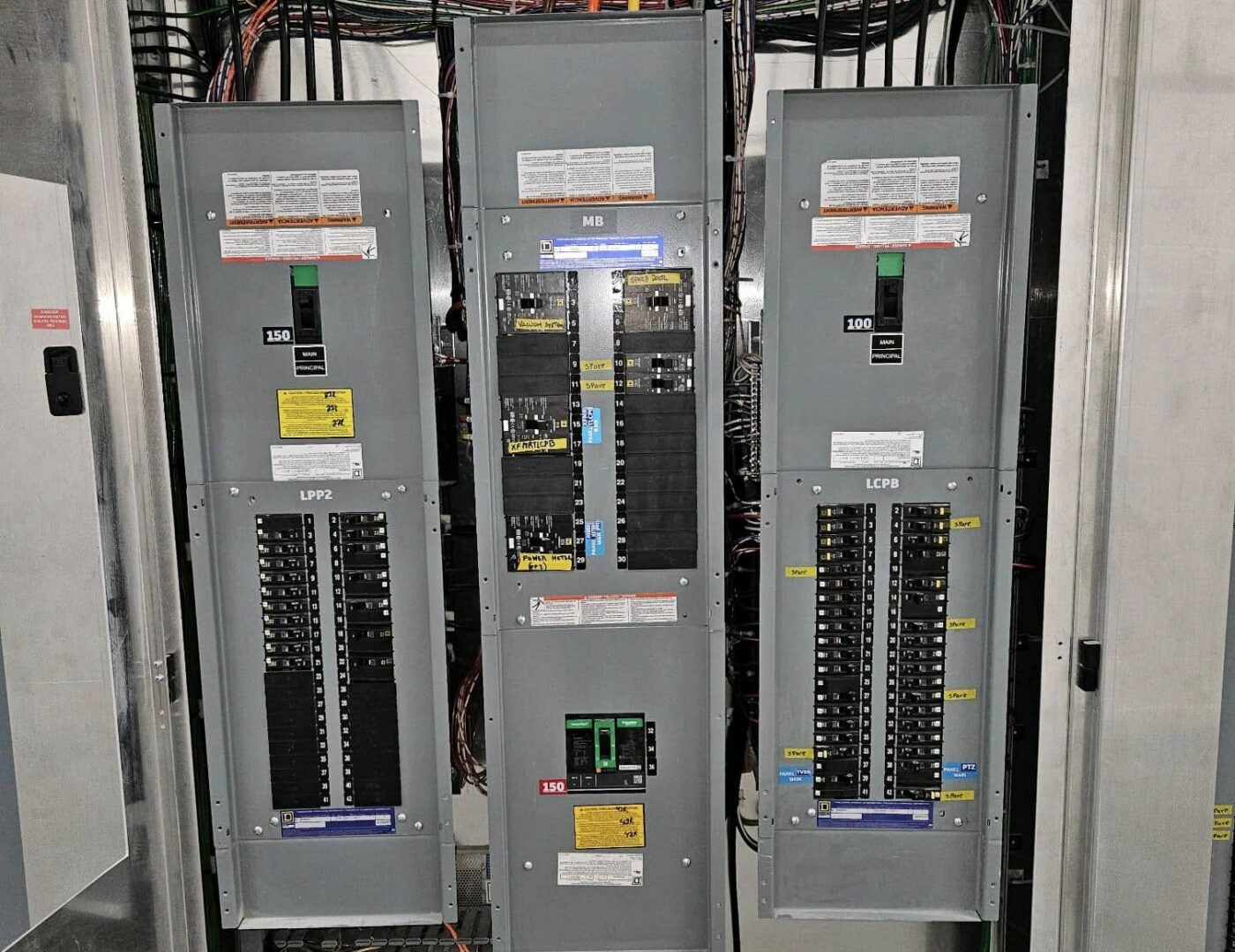 Three electrical panels with labeled circuit breakers sit side by side, colorful wires above and behind them in an industrial setting.