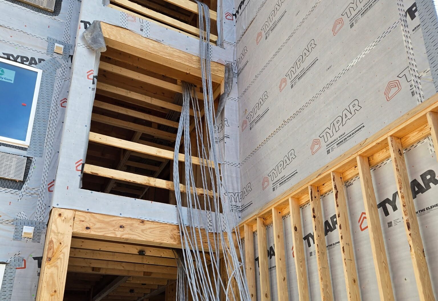 A building being constructed with visible wooden framing, Typar house wrap outside, and long electrical conduits hanging to ground level.