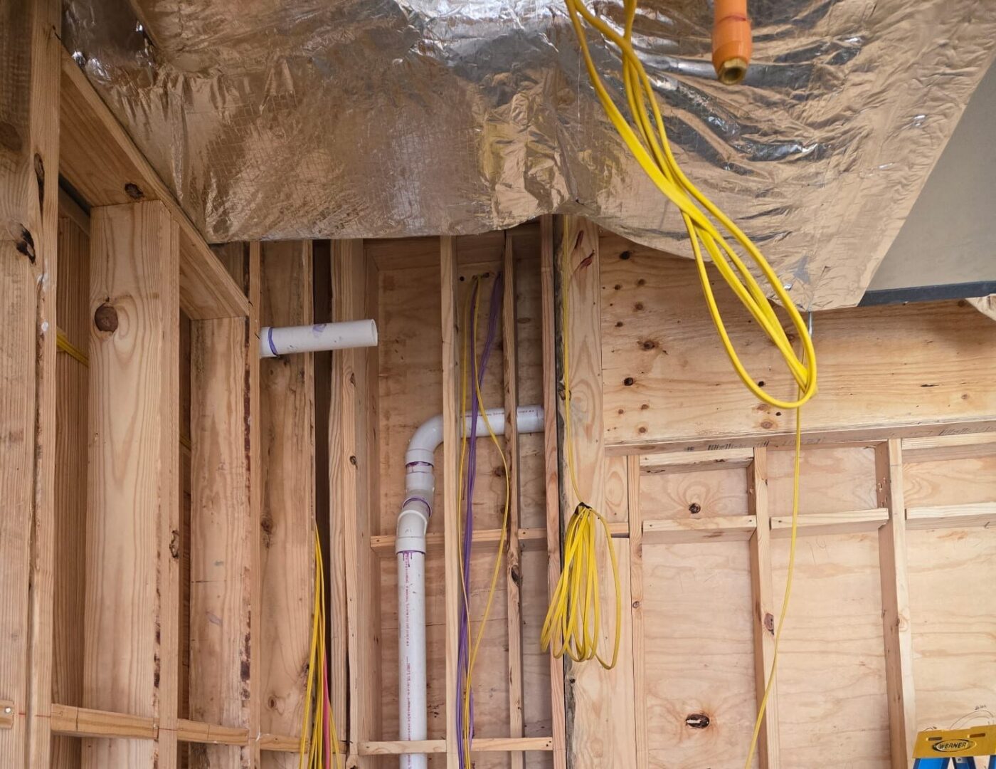 Partially built wooden wall, visible plumbing and wiring. Yellow and purple wires hang, insulation on ceiling, blue stepladder right.