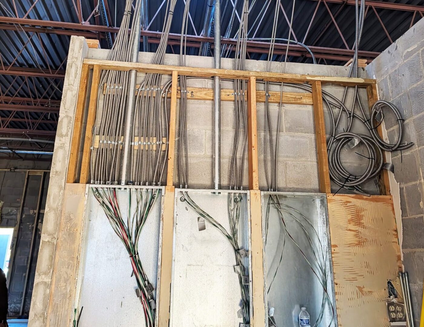 A construction site wall with exposed wiring and conduit. Unfinished ceiling with steel beams. Cables hang into open electrical panels.