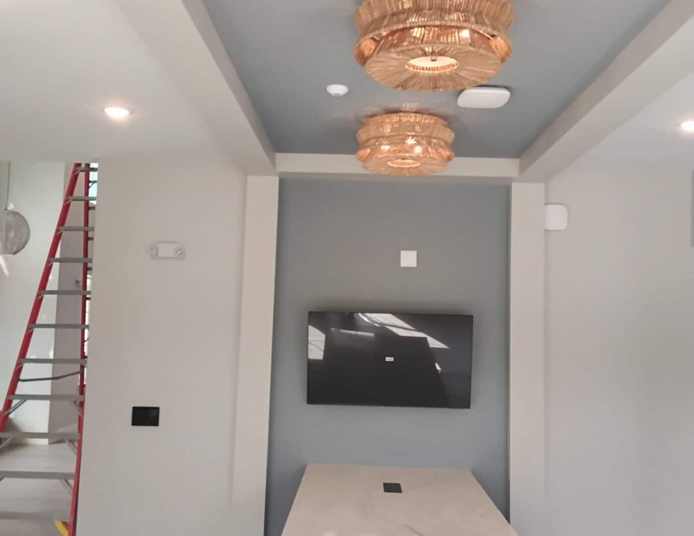 A modern interior with gray walls, marble island counter, two gold circular ceiling lights, wall-mounted TV, and red ladder by stairs.