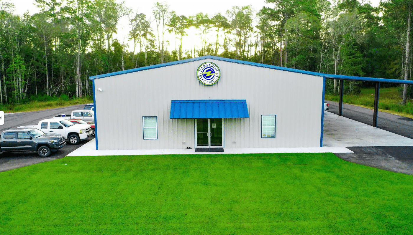 A single-story metal building with a blue awning and circular logo above the entrance, green grass, parked cars, wooded area nearby.