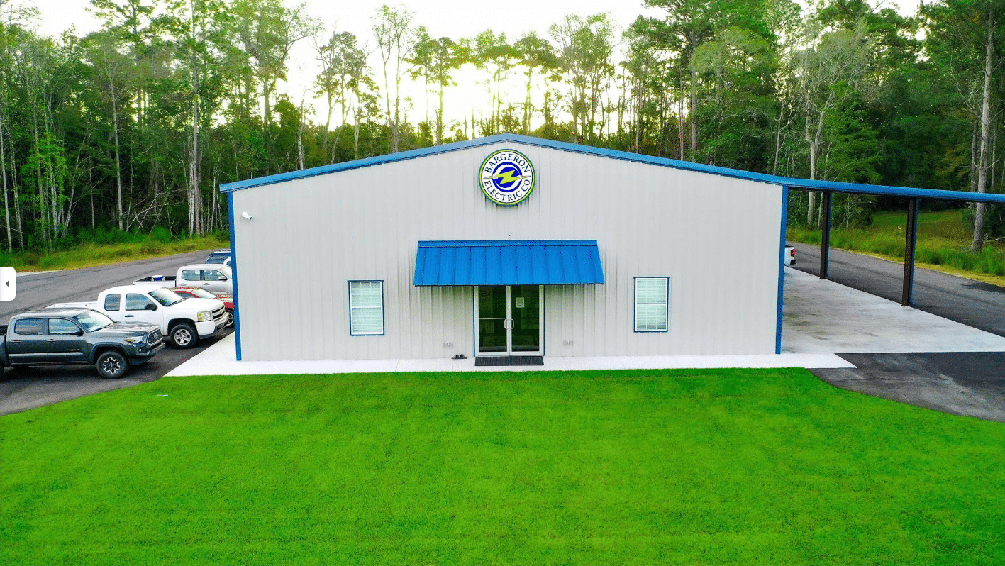 A single-story metal building with a blue awning and circular logo above the entrance, green grass, parked cars, wooded area nearby.