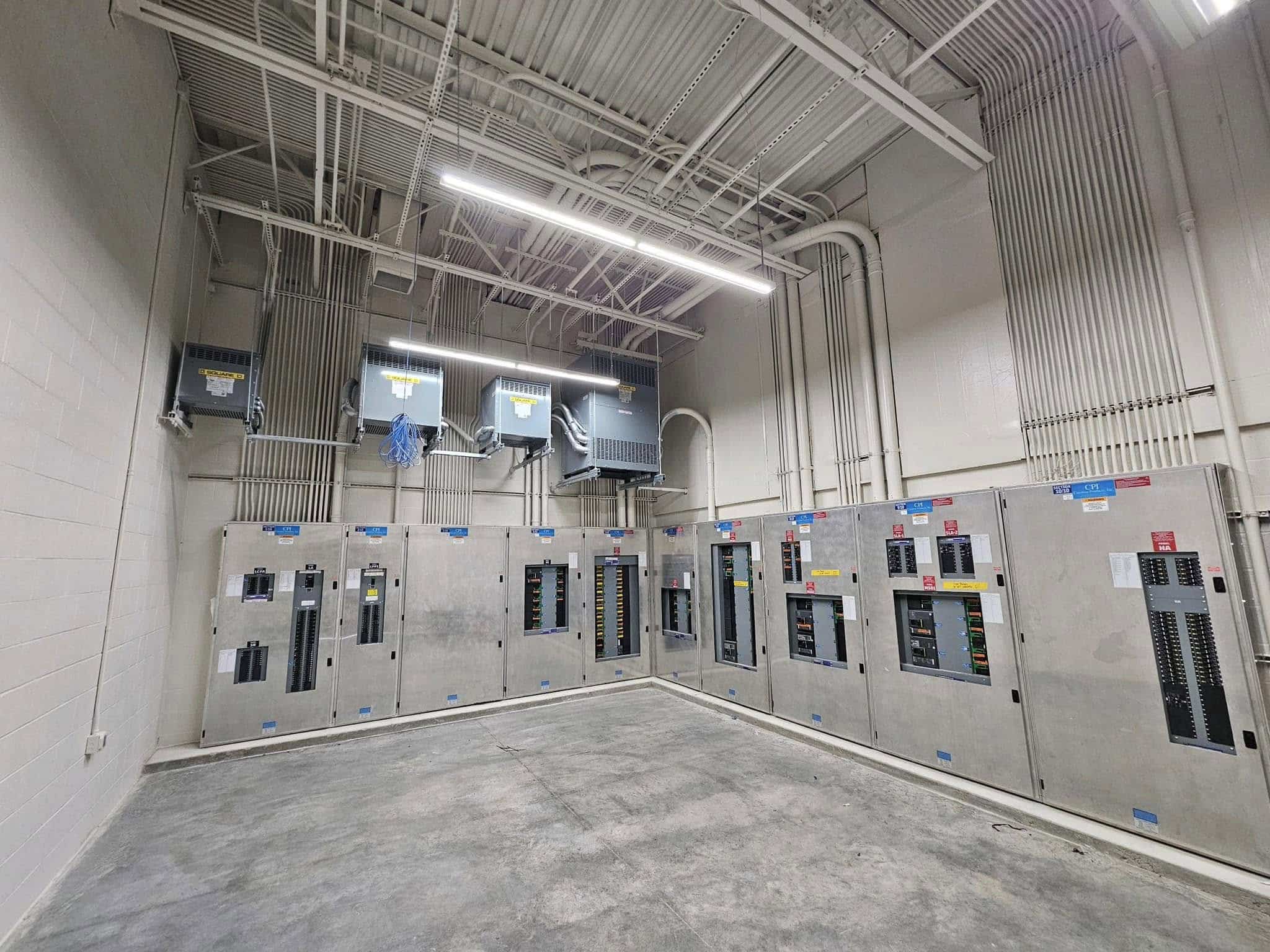 A room with large metal electrical panels on two walls, many switches, conduits overhead, unfinished concrete floor, and bright LED lighting.