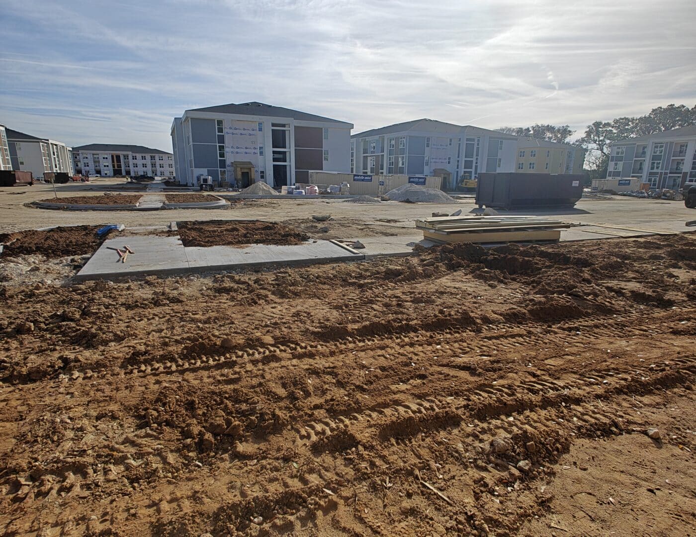 A construction site with dirt tracks and concrete foundations in front, and several apartment buildings under a sunny sky behind.
