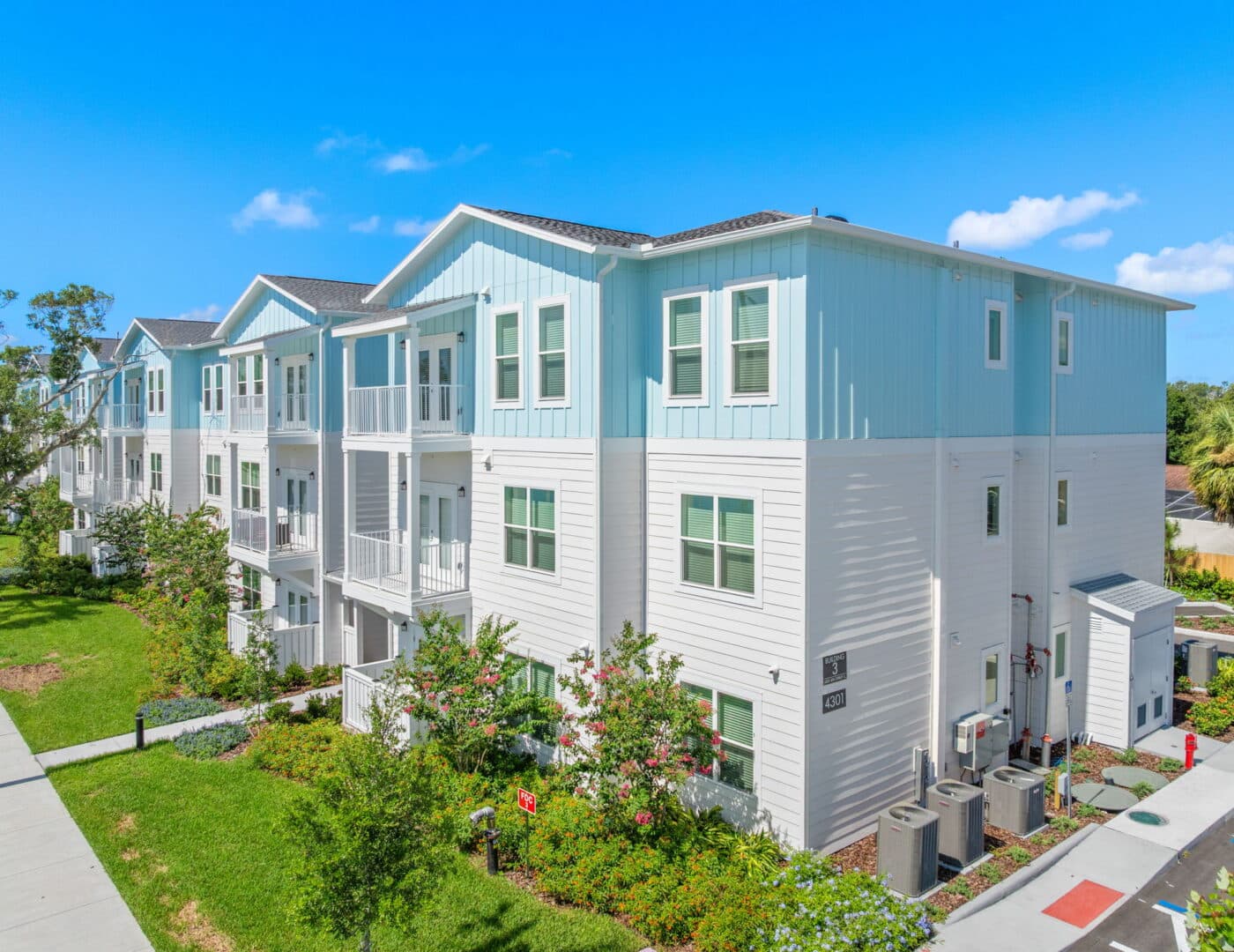Modern three-story blue and white apartments in a row, green lawns and landscaping, set under a bright sky with scattered clouds.