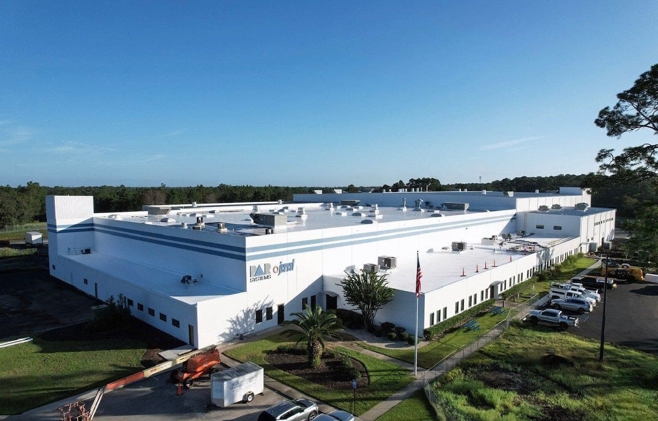 A large white industrial building with blue trim and the Oxford Science logo, surrounded by trees and parked cars under a clear sky.