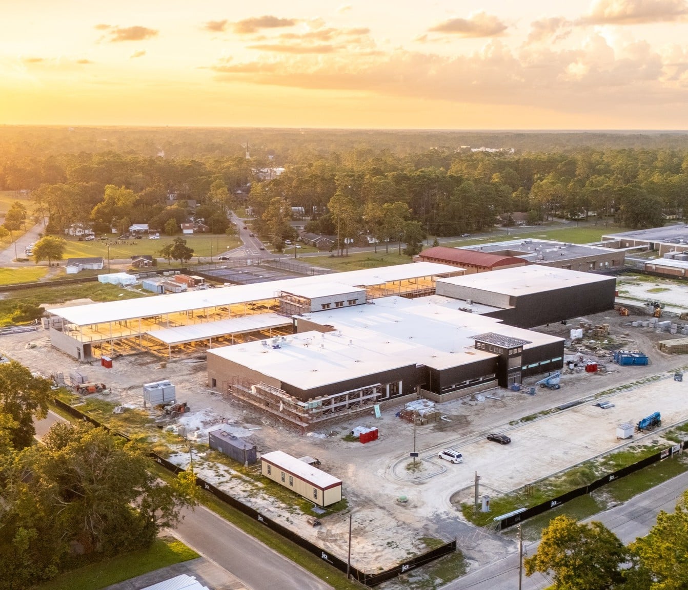 Aerial view of a large building under construction at sunset, surrounded by trees, roads, other structures, vehicles, and materials.