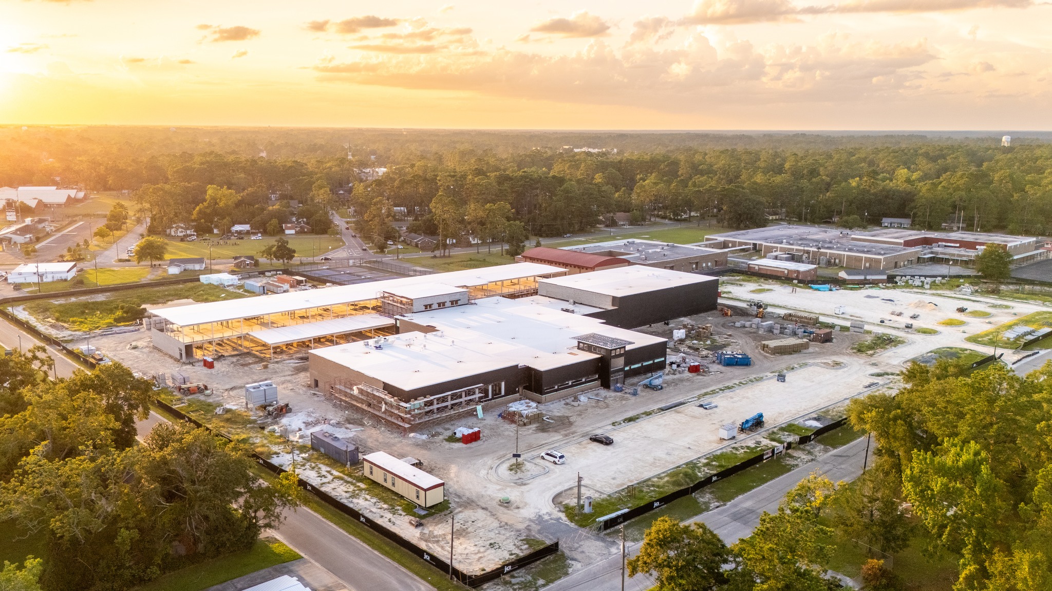 Aerial view of a large construction site amid trees and roads at sunset, with nearby buildings and homes in the background.