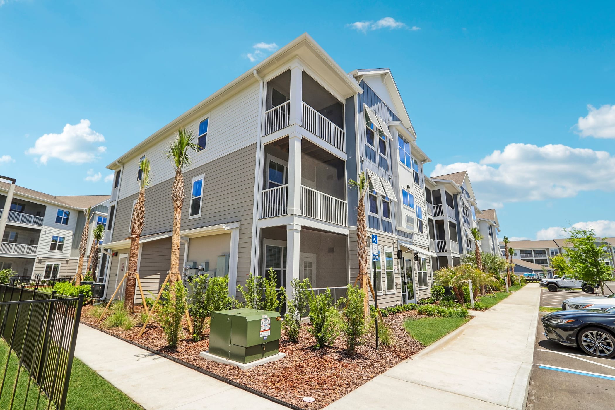 Modern three-story apartment building with balconies, gray siding, and white trim. Palm trees and cars under a blue sky.