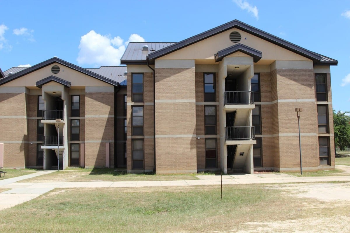 Three-story tan brick apartment building featuring small balconies and an external staircase, on grass beneath a blue sky.
