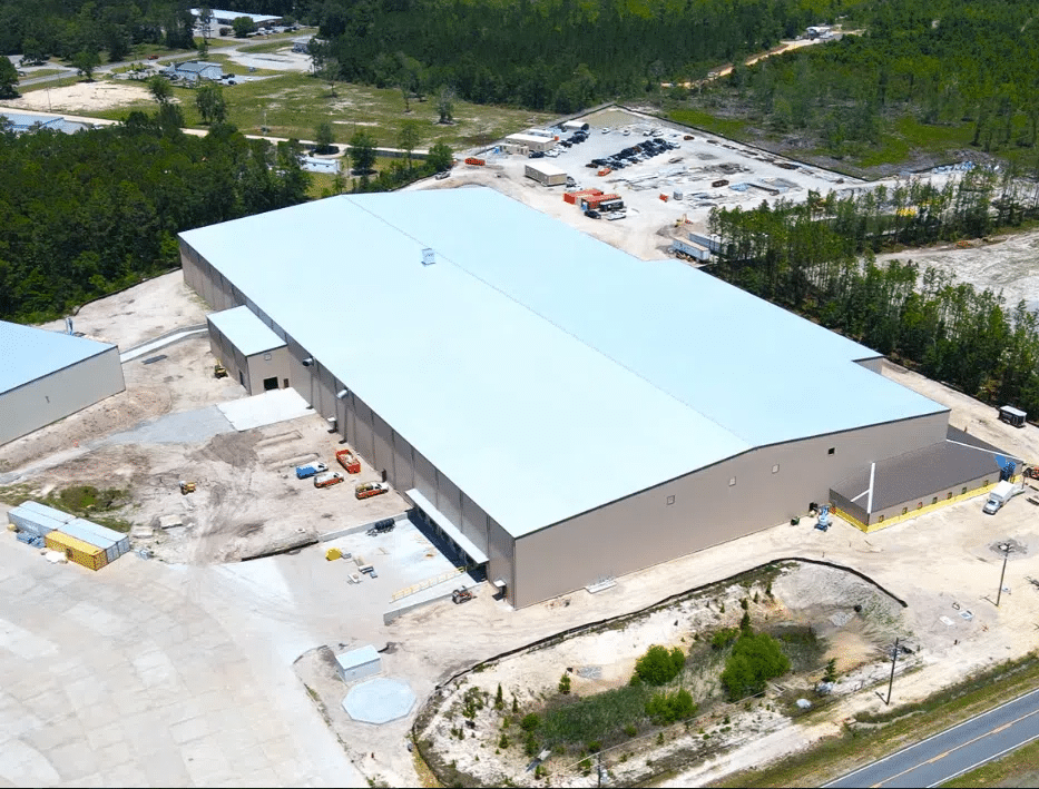 A large industrial warehouse with a white roof is surrounded by construction equipment and materials in a clearing near a road.