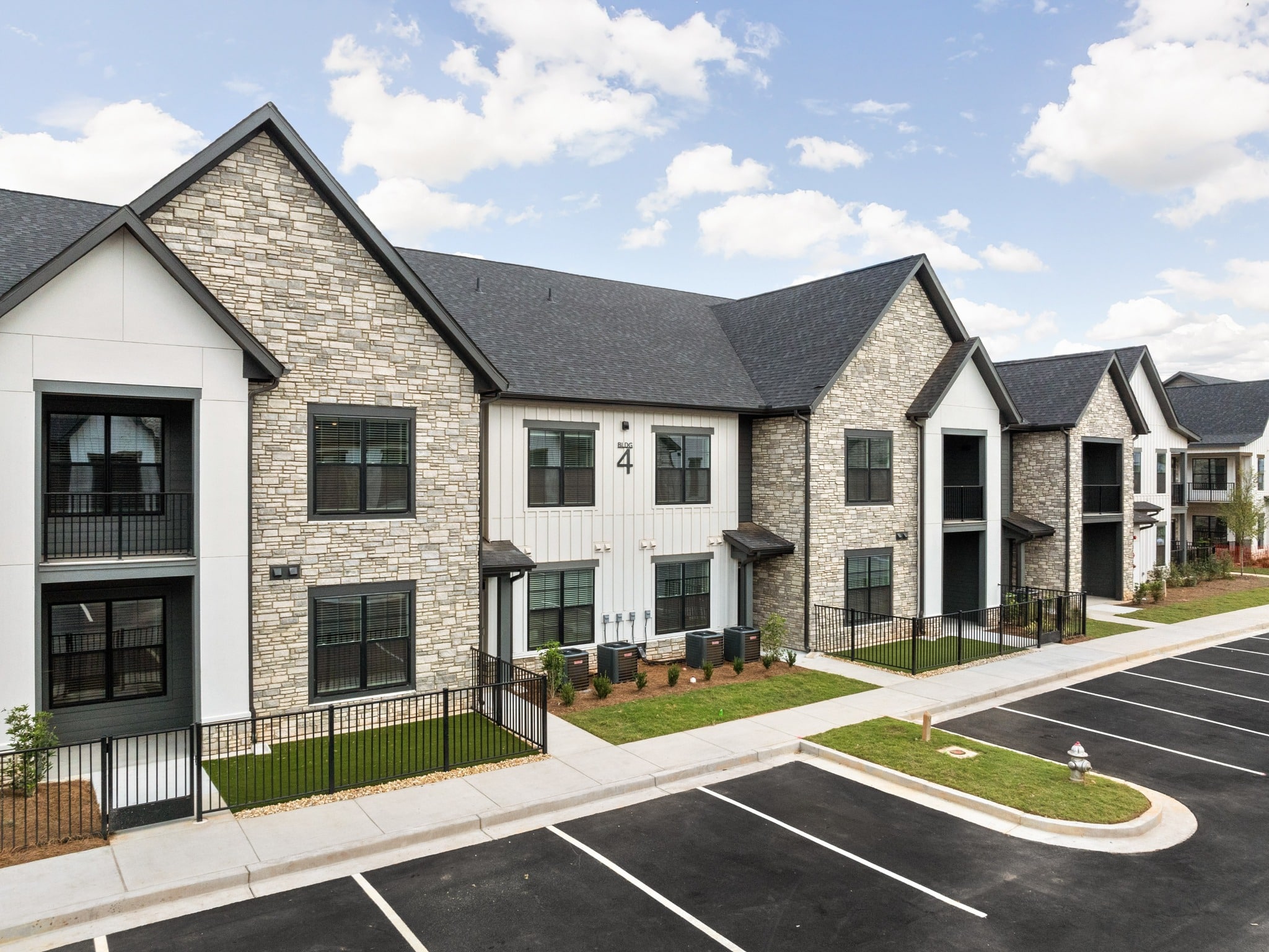 Modern apartment with stone and white siding, black trim, balconies, lawn, black fencing, empty parking lot under blue sky.