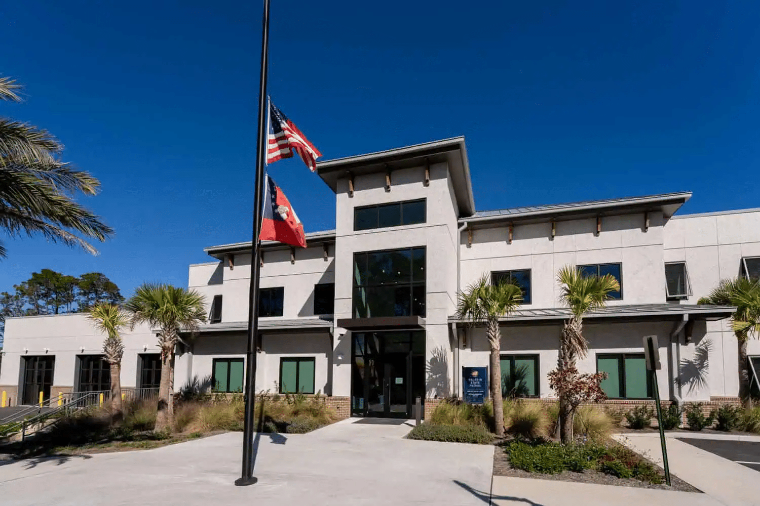 Modern two-story building with large windows, palm trees, and flags at half-staff, set against a clear blue sky.